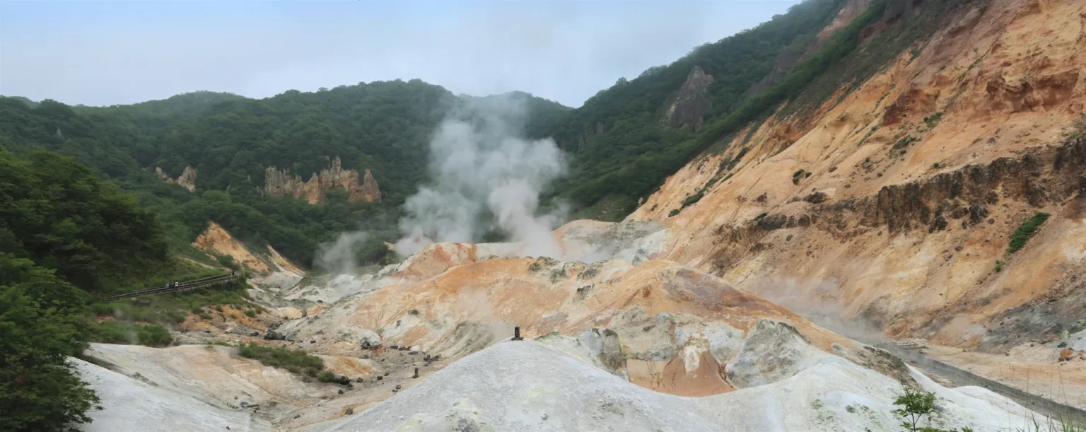 Panorama of Jigokudani in Noboribetsu in summer: green forested hills around a crater floor of white, grey and orange volcanic rock, a large plume of steam rising in the centre and a wooden walkway on the left edge