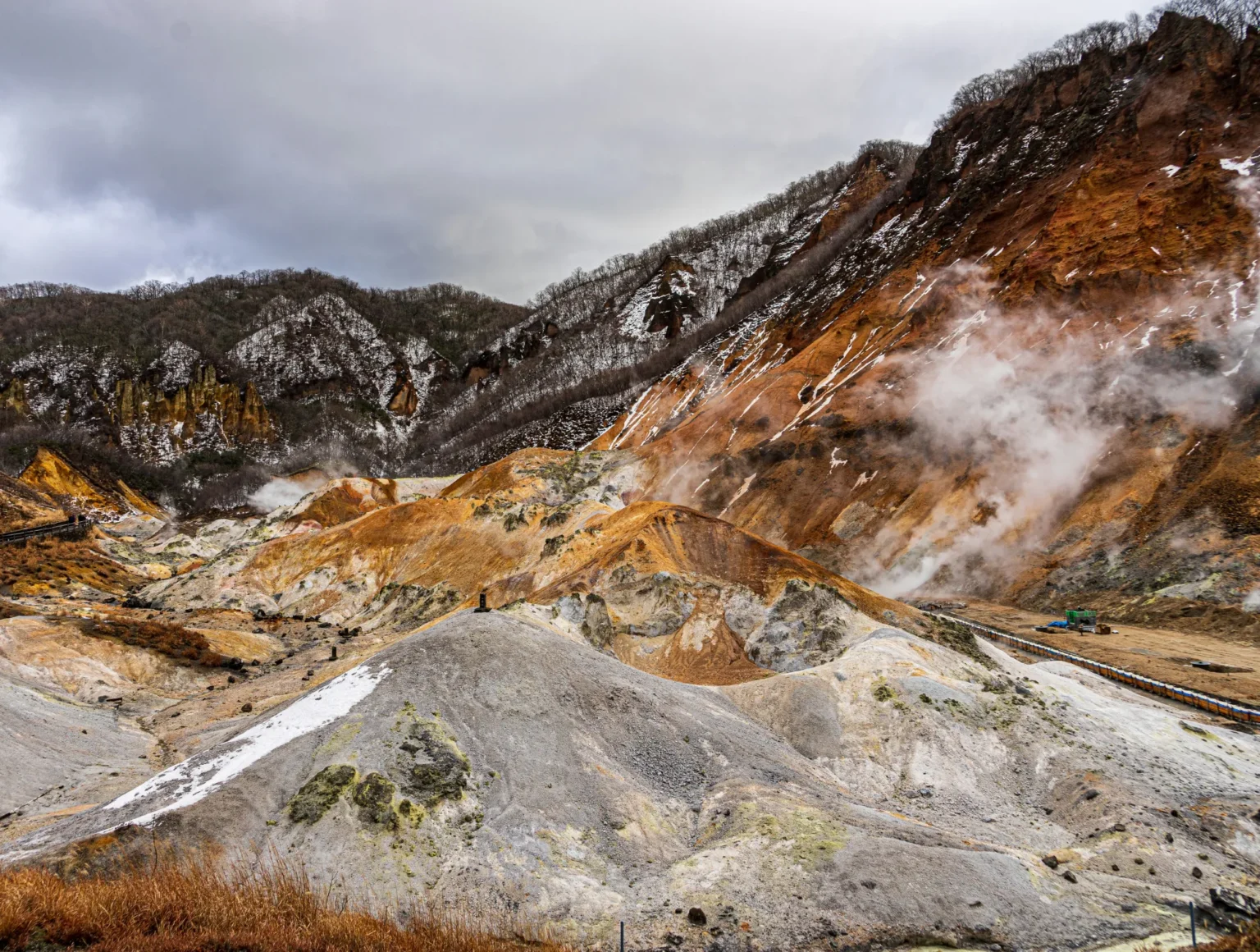 Jigokudani in Noboribetsu under grey cloud: grey and orange mounds of volcanic rock with steam drifting