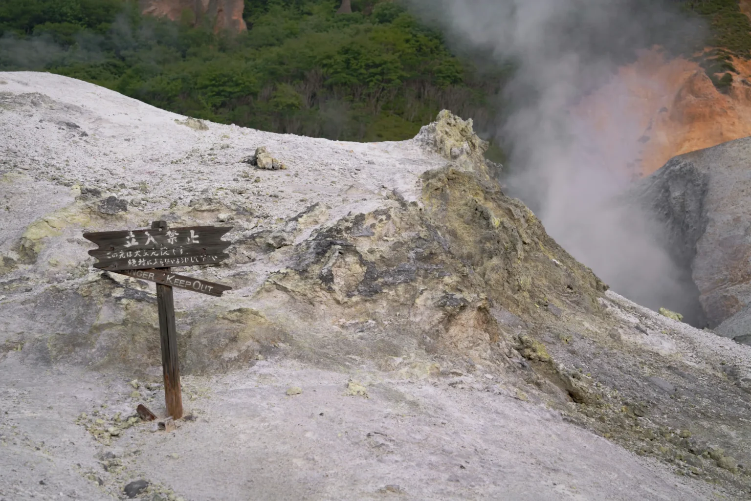 A weathered wooden sign on pale volcanic rock in Noboribetsu reading, in Japanese, no entry, very dangerous beyond this point, never enter, with a lower board reading Danger Keep Out, beside a steaming vent and a green wooded slope
