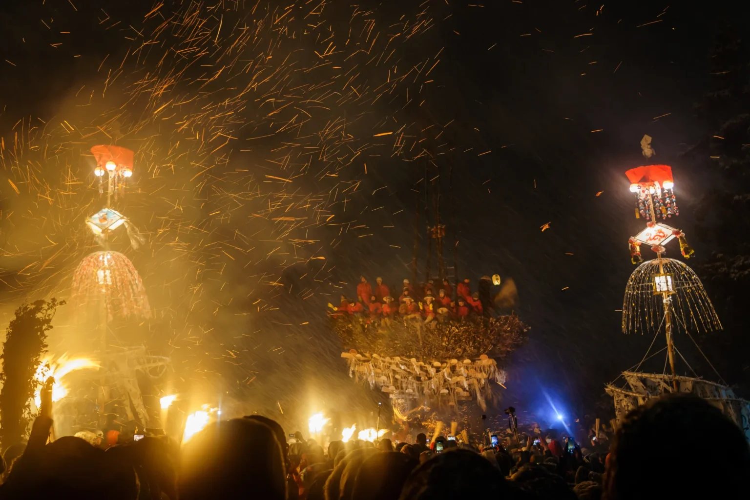At night, the wooden shaden crowded on top with men dressed in red, a tall decorated lantern pole on each side and sparks in the sky
