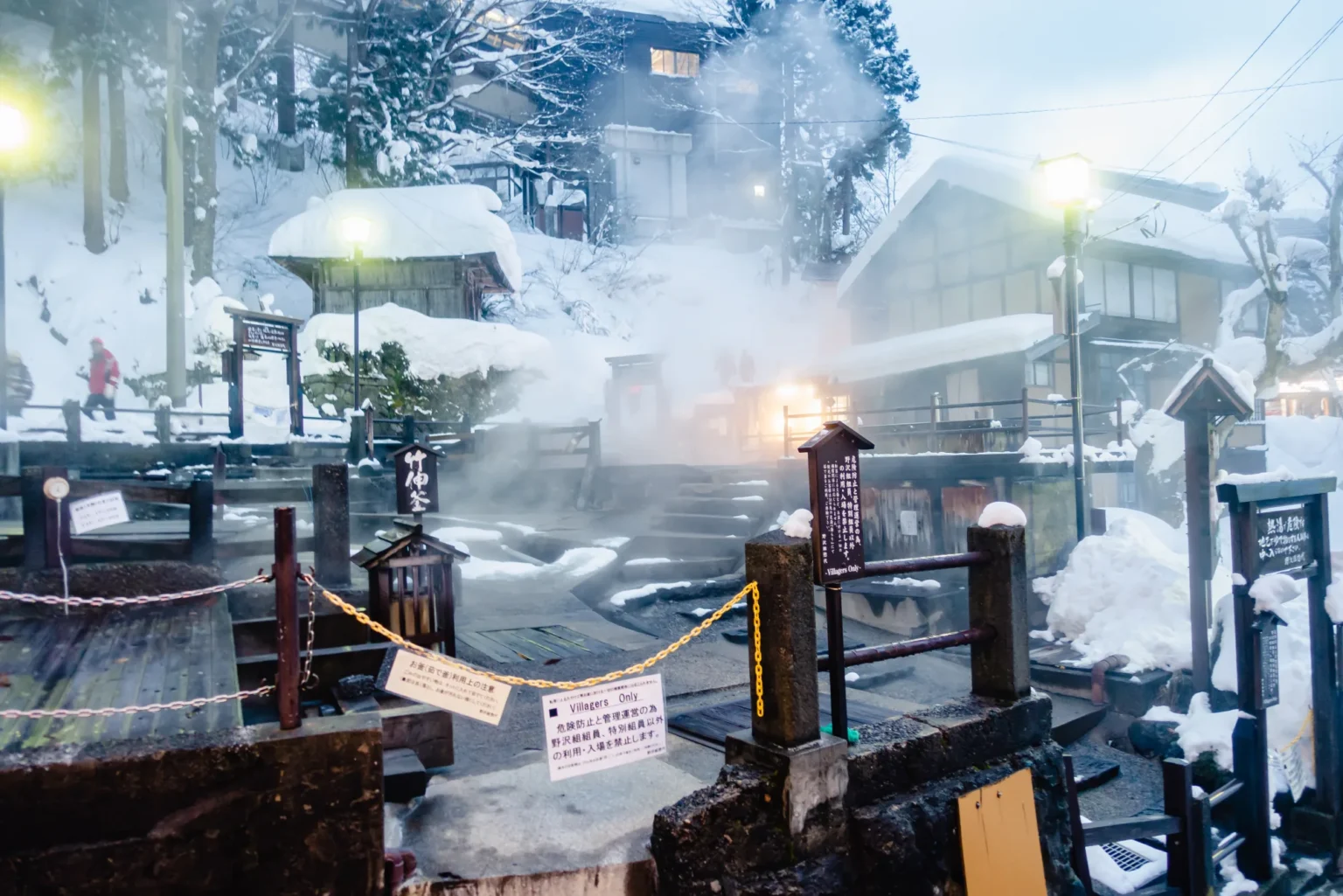 Steam rising from hot spring pools among deep snow, fenced off with a chain and rails, with a sign reading Villagers Only