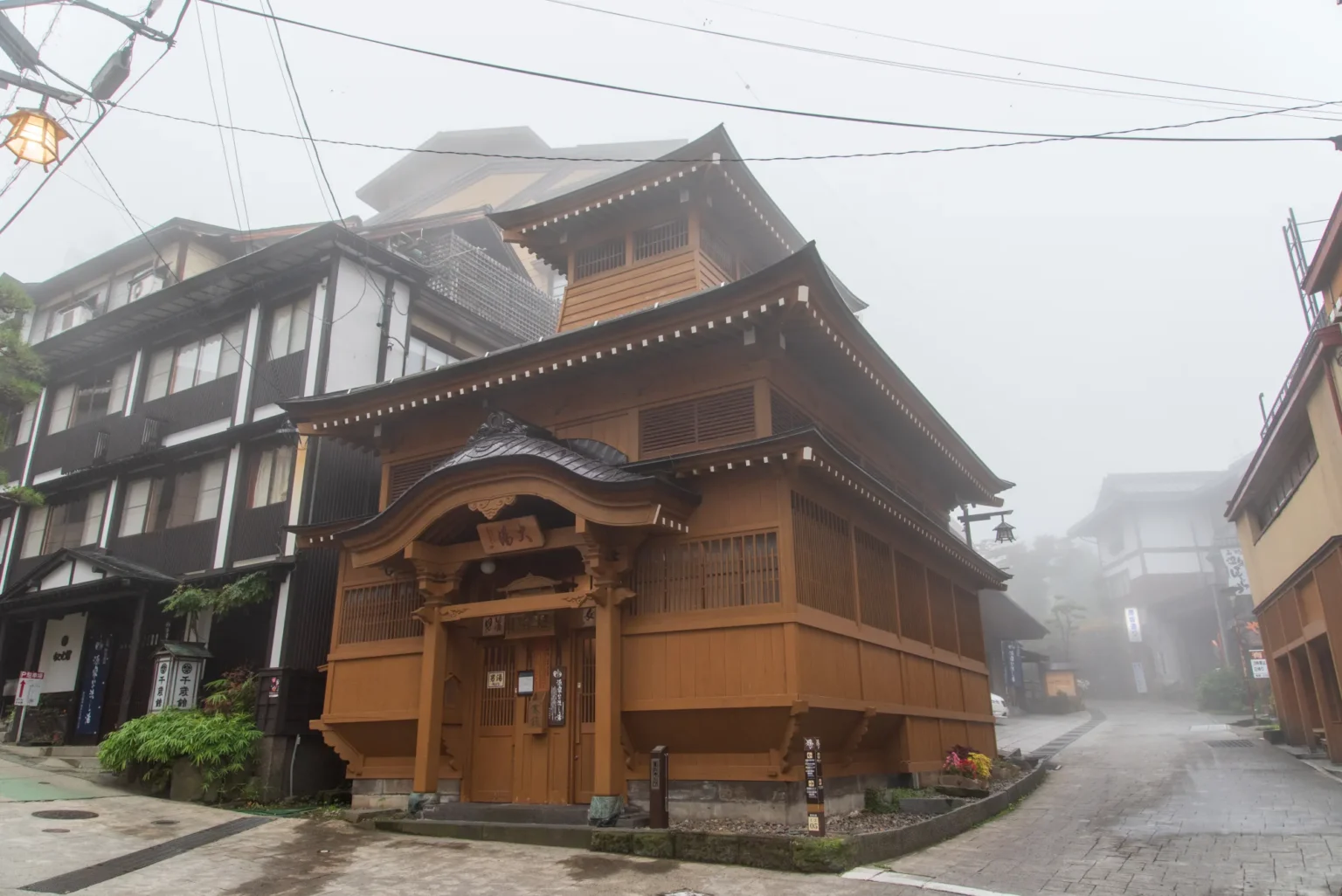 The wooden Oyu public bathhouse in Nozawa Onsen, with layered tiled roofs and a small tower, at a corner of a wet stone-paved street on a misty day, inns on either side