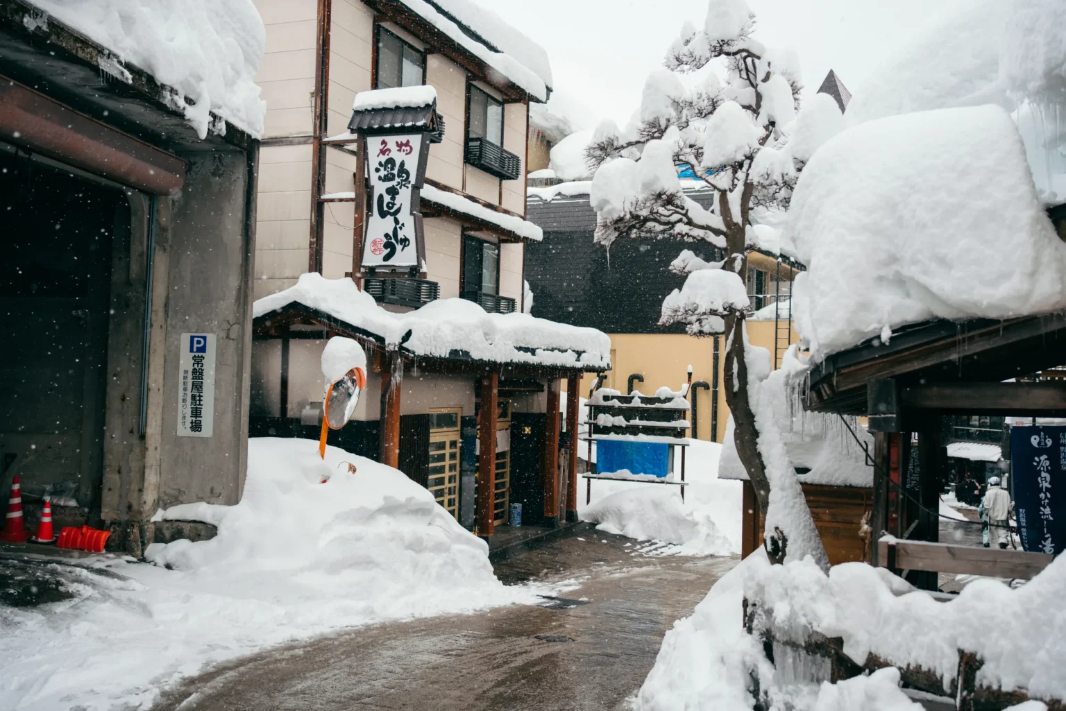 Snow falling on a narrow street in Nozawa Onsen, with deep snow banked against the buildings, a hanging shop sign for onsen manjū buns, a private parking sign and a snow-laden pine