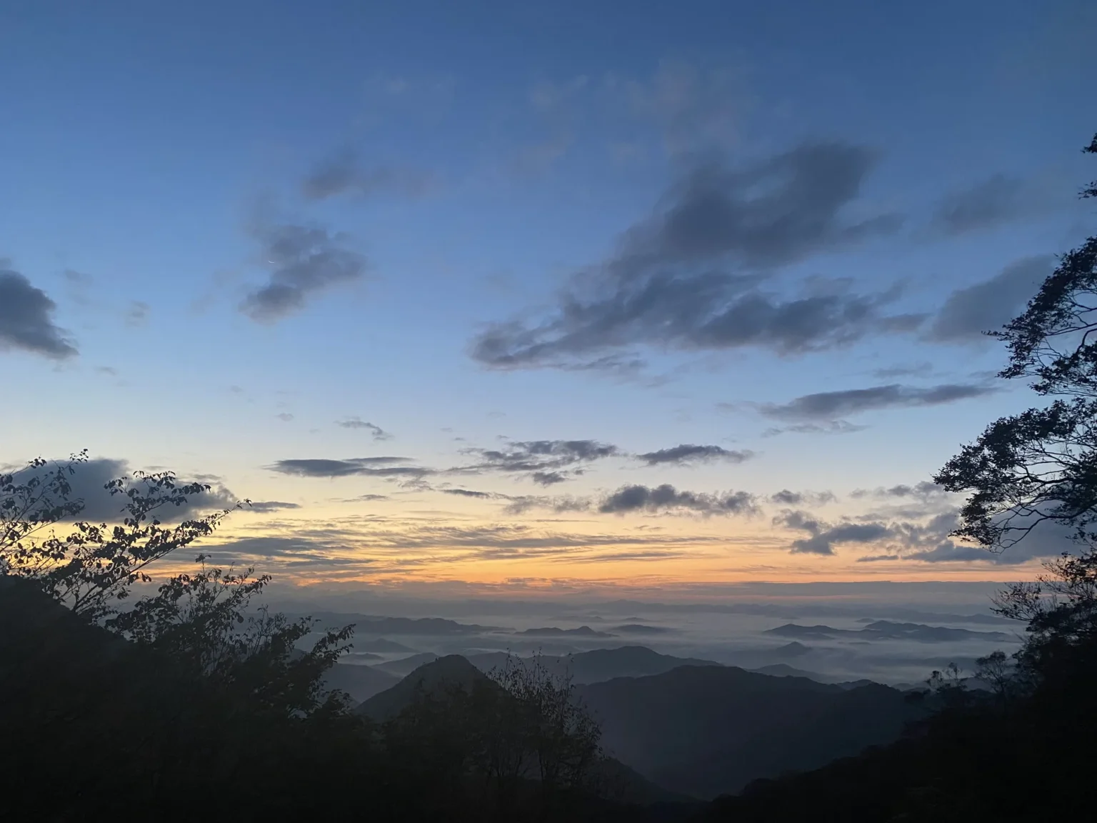 Blue sky with grey clouds before sunrise, an orange band along the horizon, and white fog lying in the valleys between dark forested ridges, photographed at 6:01 a.m. on Mount Oe, Fukuchiyama, on 3 November 2021