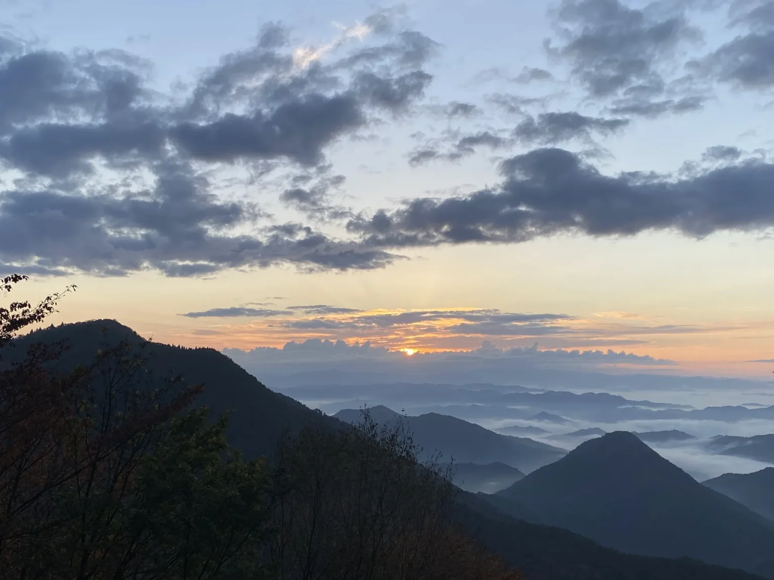 The sun rising over a bank of cloud on the horizon at 6:31 a.m., with white fog filling the valleys between dark forested ridges and a pointed peak standing out of it, seen from Mount Oe in Fukuchiyama, Kyoto Prefecture, on 3 November 2021