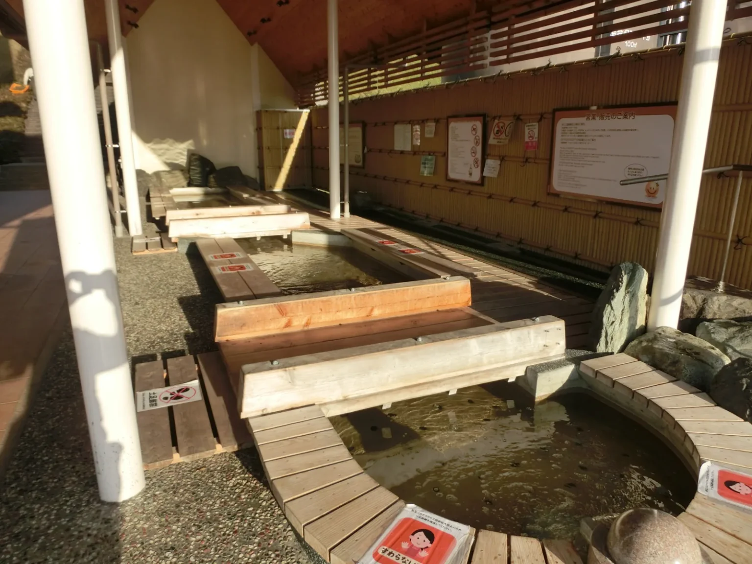 A covered foot bath with wooden edges and benches around shallow pools of water, signs on a bamboo-lined wall behind, at Ogoto Onsen Tourism Park