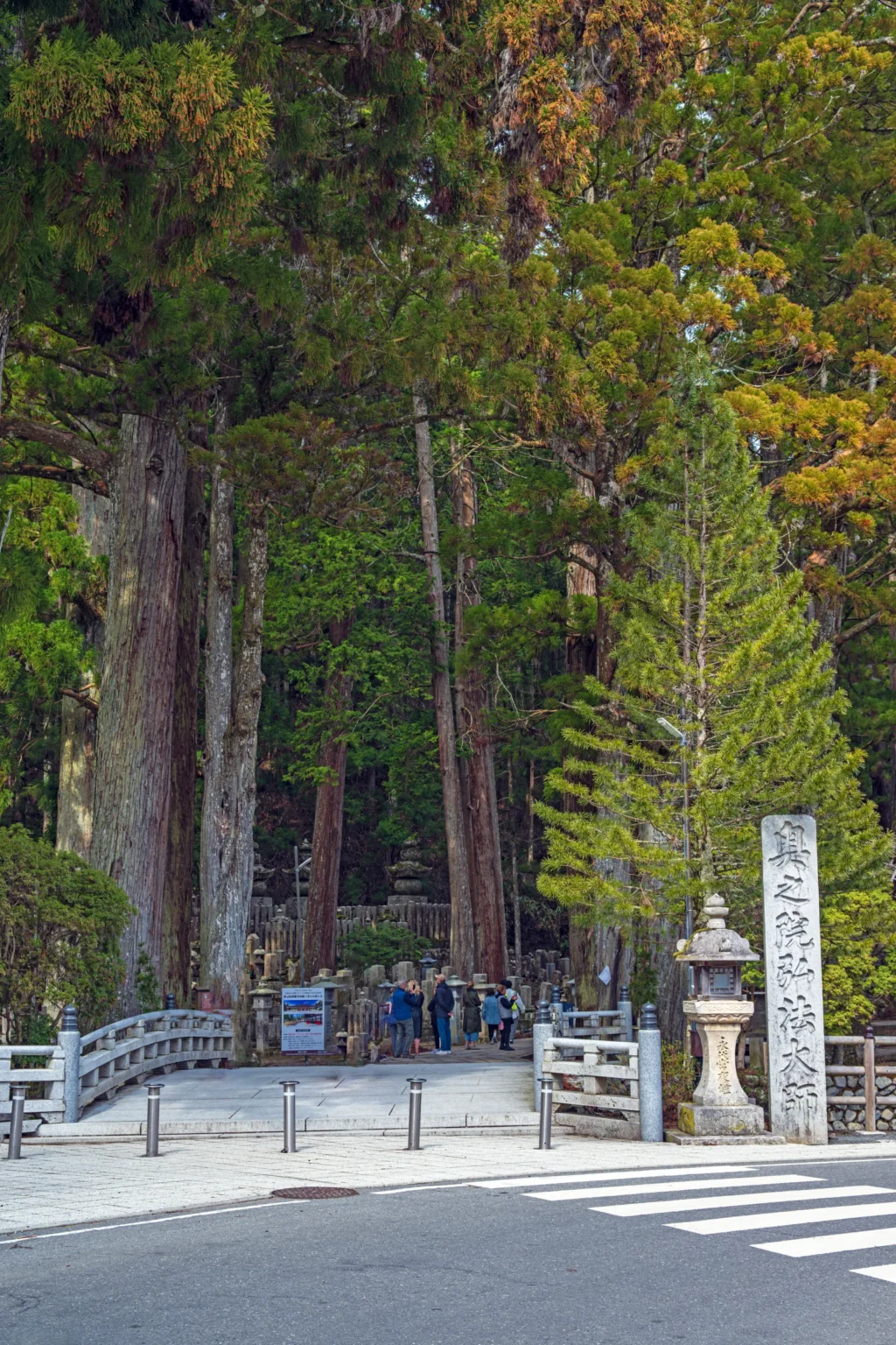The entrance to the Okunoin approach at Ichinohashi, with a small bridge, a stone pillar and tall cedar trees above the road