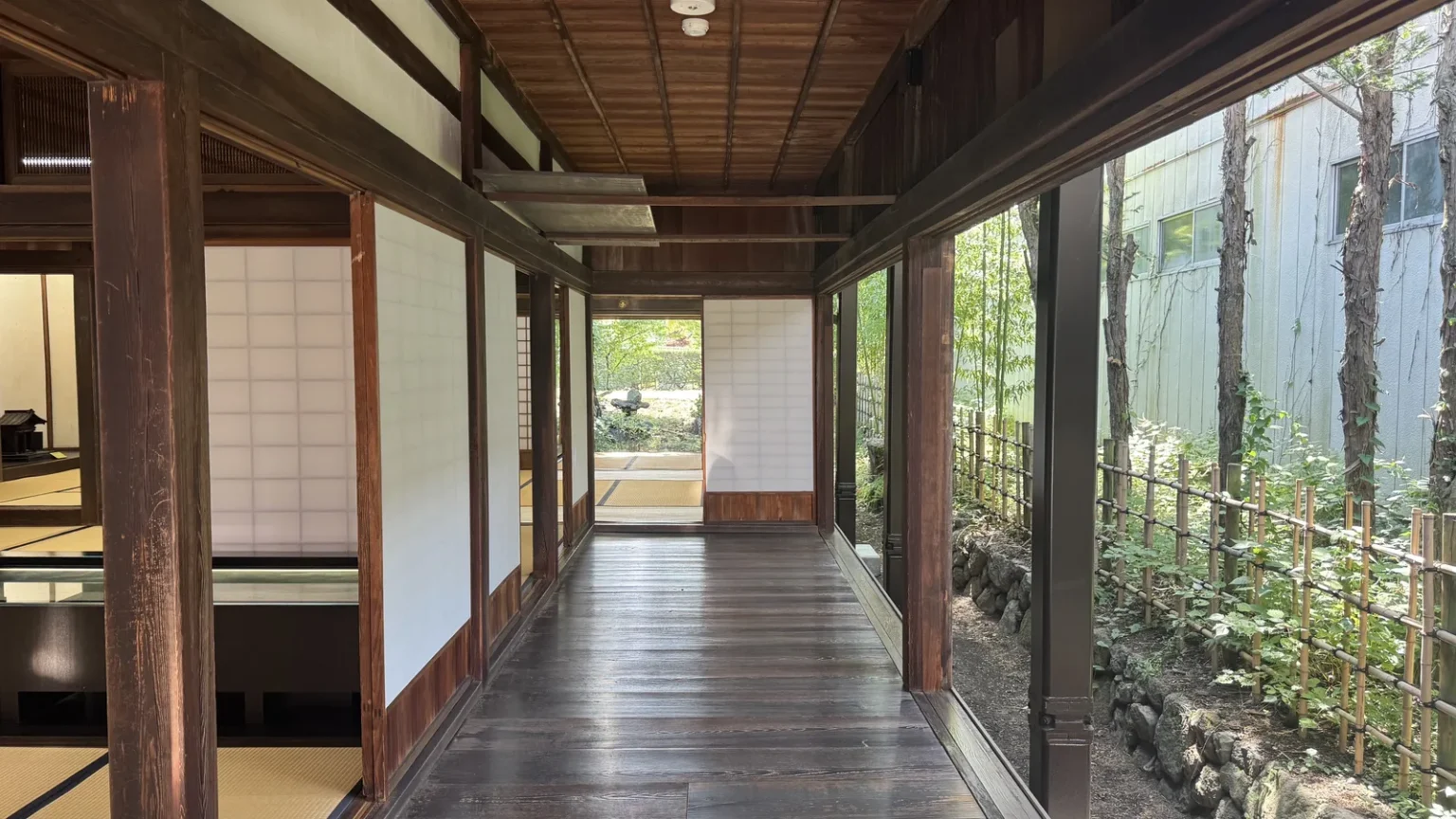 A polished wooden corridor running between paper screens on the left and glass windows onto greenery on the right