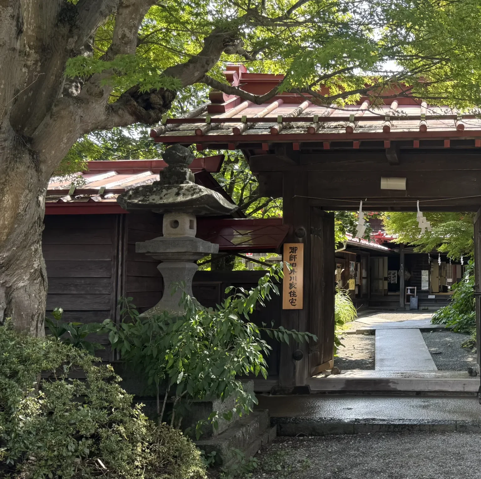 A roofed wooden gate with red roof trim and a stone lantern, opening onto a path to the house under green leaves