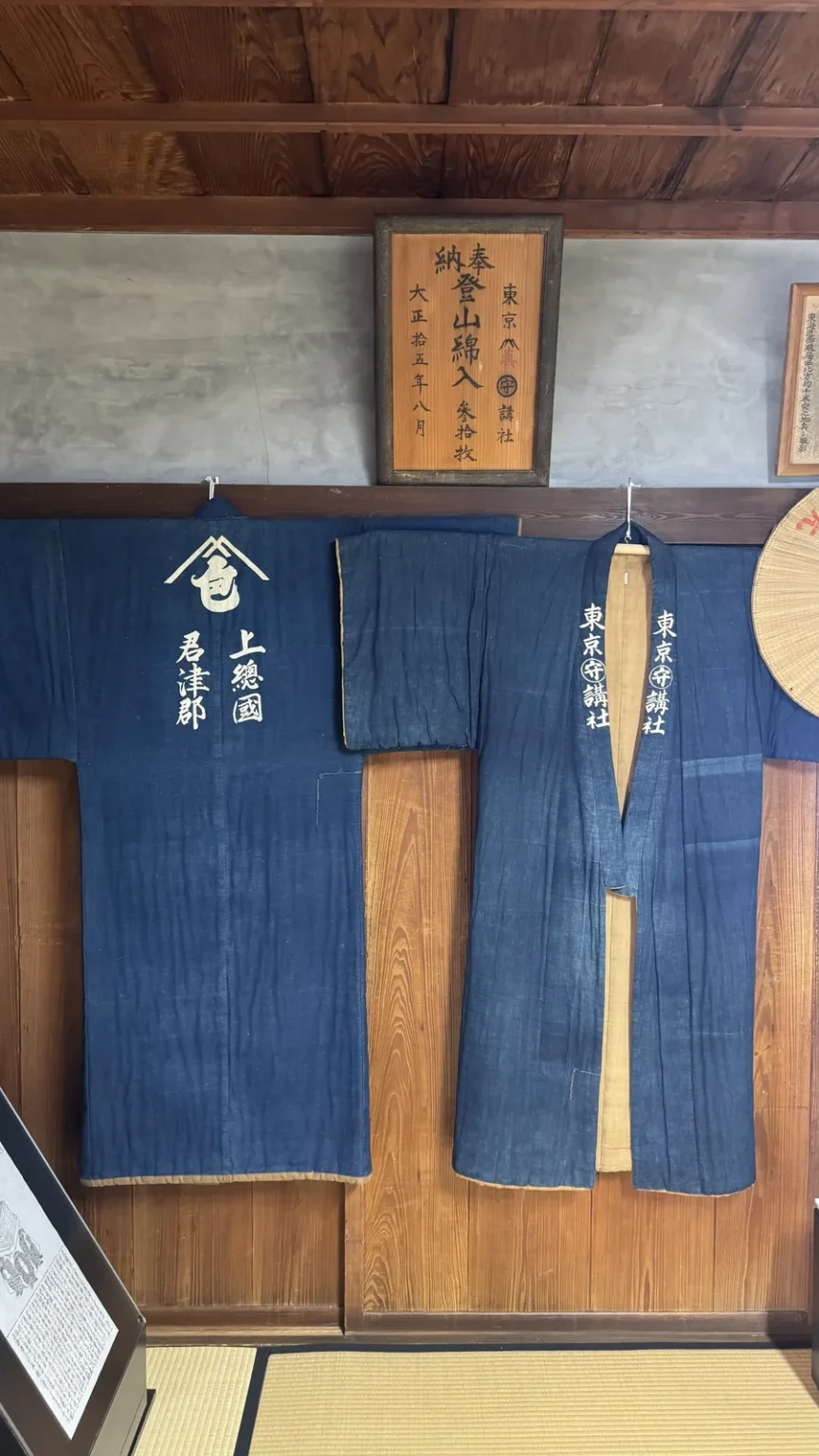 Two indigo jackets with white Japanese lettering hanging on a wooden wall below a framed wooden plaque
