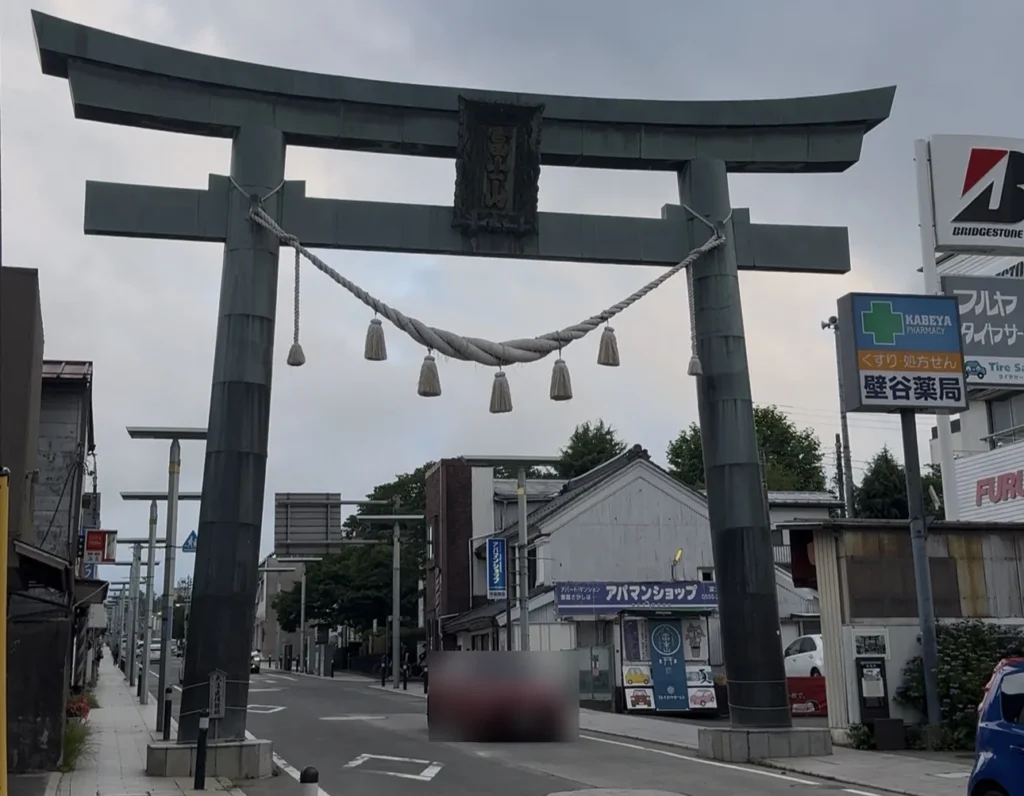 A large copper-green torii with a thick rope and tassels spanning a town street, with a car, blurred, passing underneath