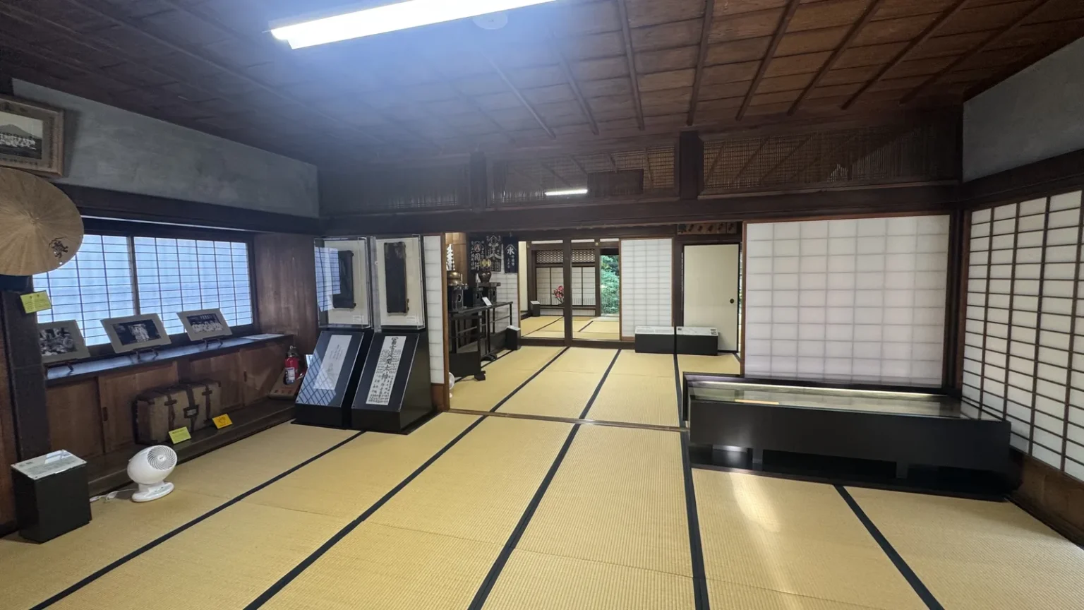 A long tatami hall under a dark wooden ceiling, with display cases along the left wall and paper sliding doors on the right