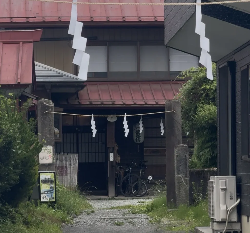 A house entrance with a rope and zigzag white paper streamers across it, red roofs, a gravel path and a bicycle under the eaves
