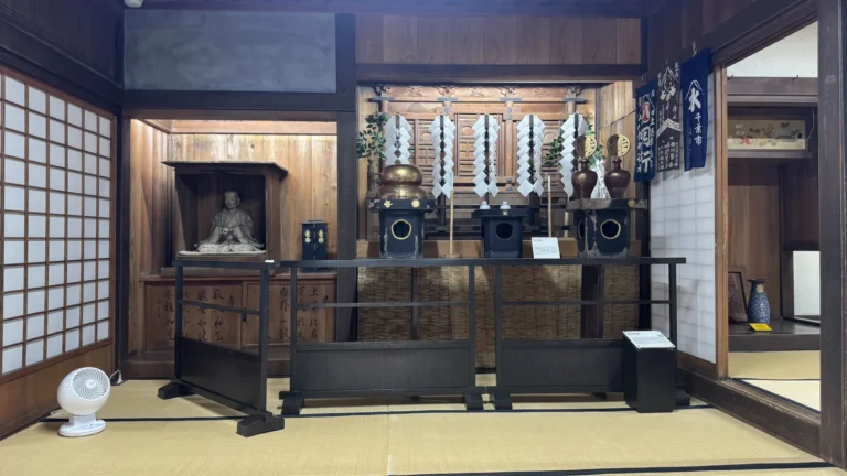 A tatami room with a wooden altar holding a seated statue, white paper offerings on tall stands and dark lacquered vessels, behind a low wooden rail