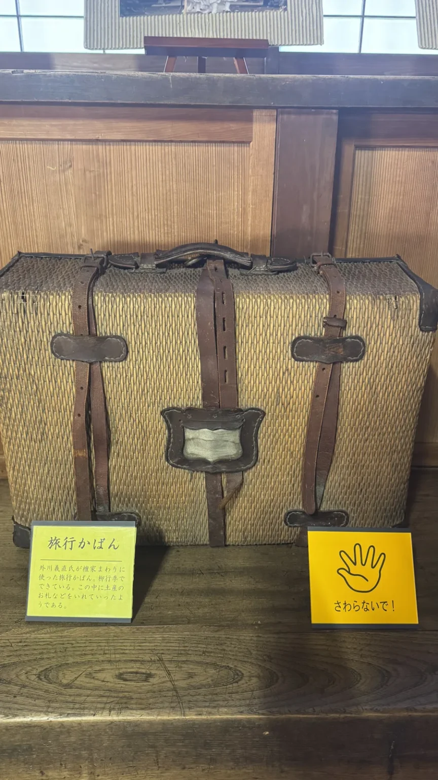 A woven wicker travel case with straps on a wooden shelf, with a Japanese label and a yellow do-not-touch sign
