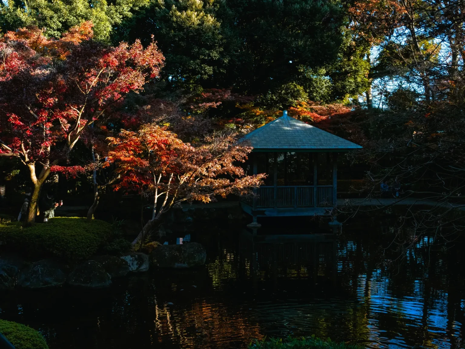 A small pavilion with a grey pyramid roof on the edge of a dark pond, with red and orange maples on the left and their colours reflected in the water