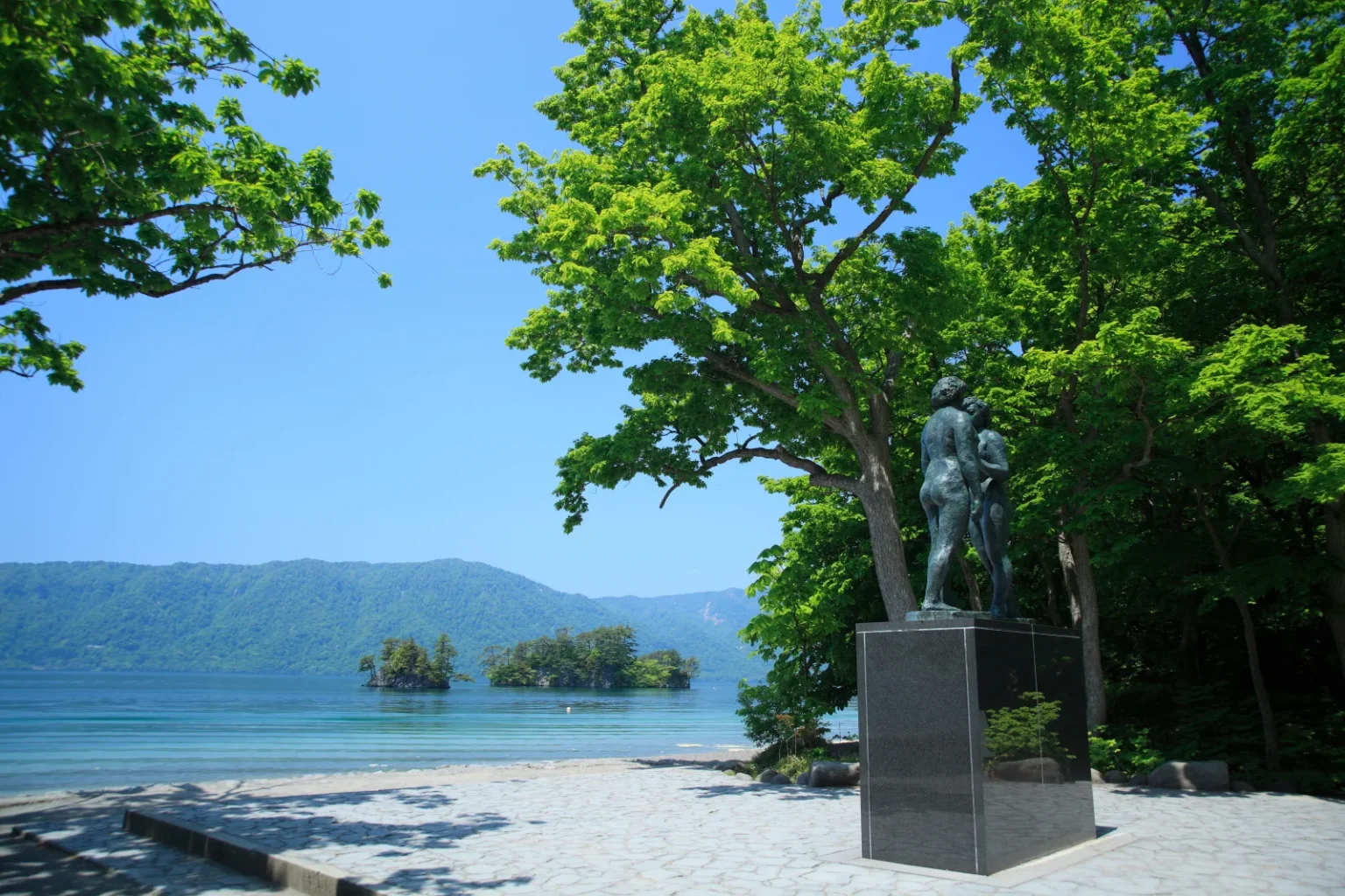 The bronze Otome statue of two figures on a dark stone base on a paved plaza by the shore of Lake Towada, with small wooded islands in the water and green trees