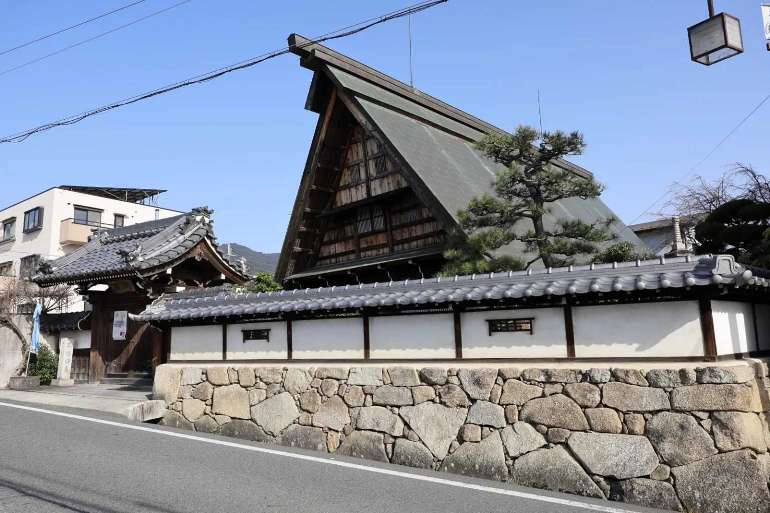 A dry-stone wall of large fitted stones topped by a white plastered wall, with a steep-roofed building behind it, in Sakamoto