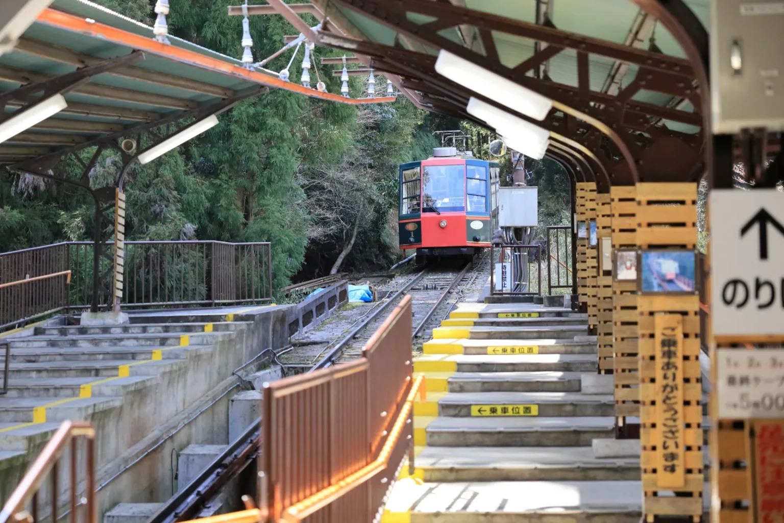 A red and cream Sakamoto Cable car waiting at the top of a stepped station platform among trees