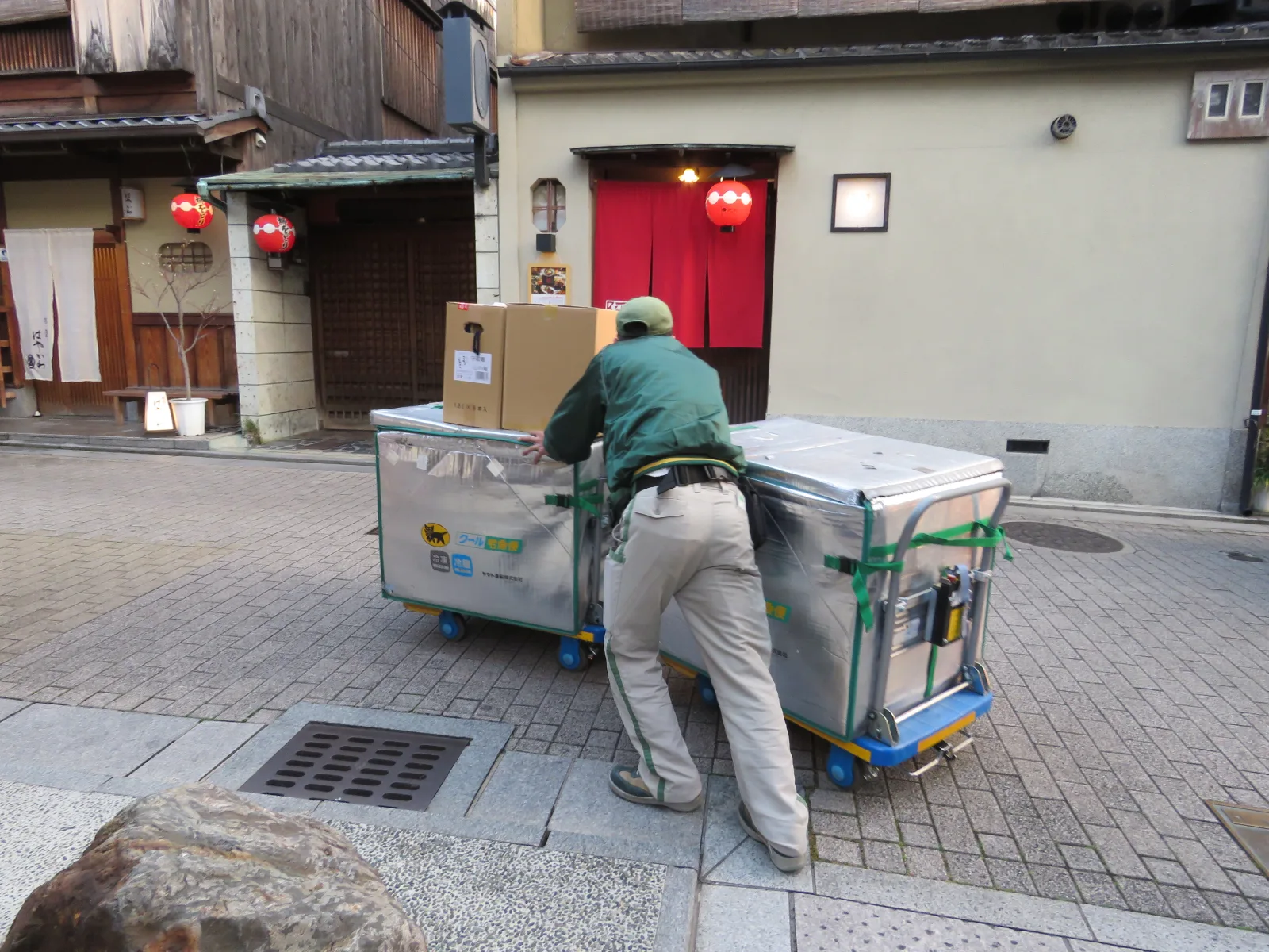 A Yamato Transport worker in a green uniform pushes two silver delivery carts with two cardboard boxes on top along a stone-paved street in Kyoto, past wooden and plastered buildings with red lanterns and a red noren curtain