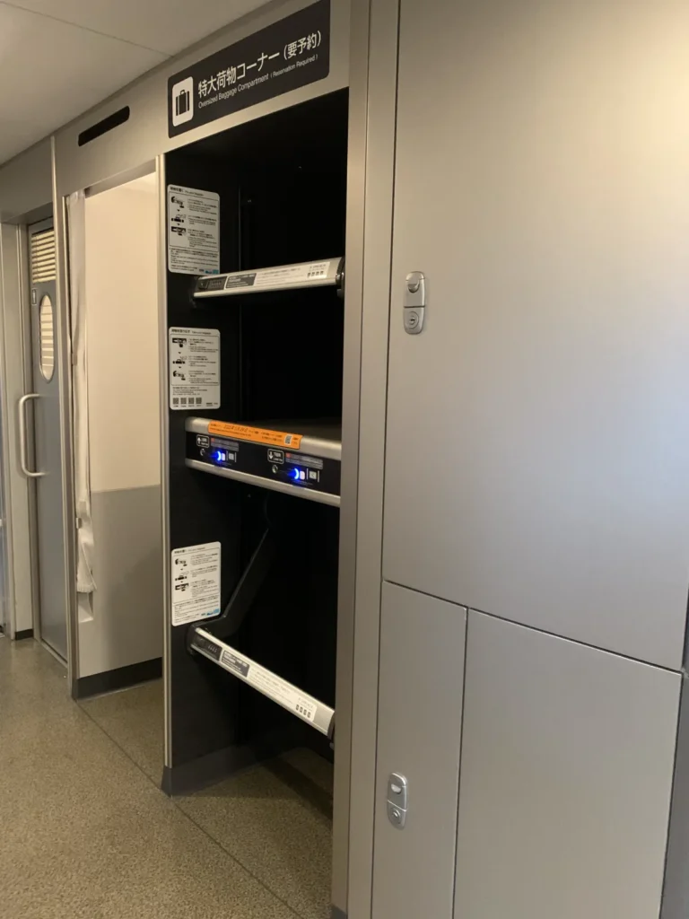 An open two-level luggage rack set into the wall of a Shinkansen deck, with bars and IC-card lock units, instruction stickers, and a sign above reading Oversized Baggage Compartment (Reservation Required) in Japanese and English