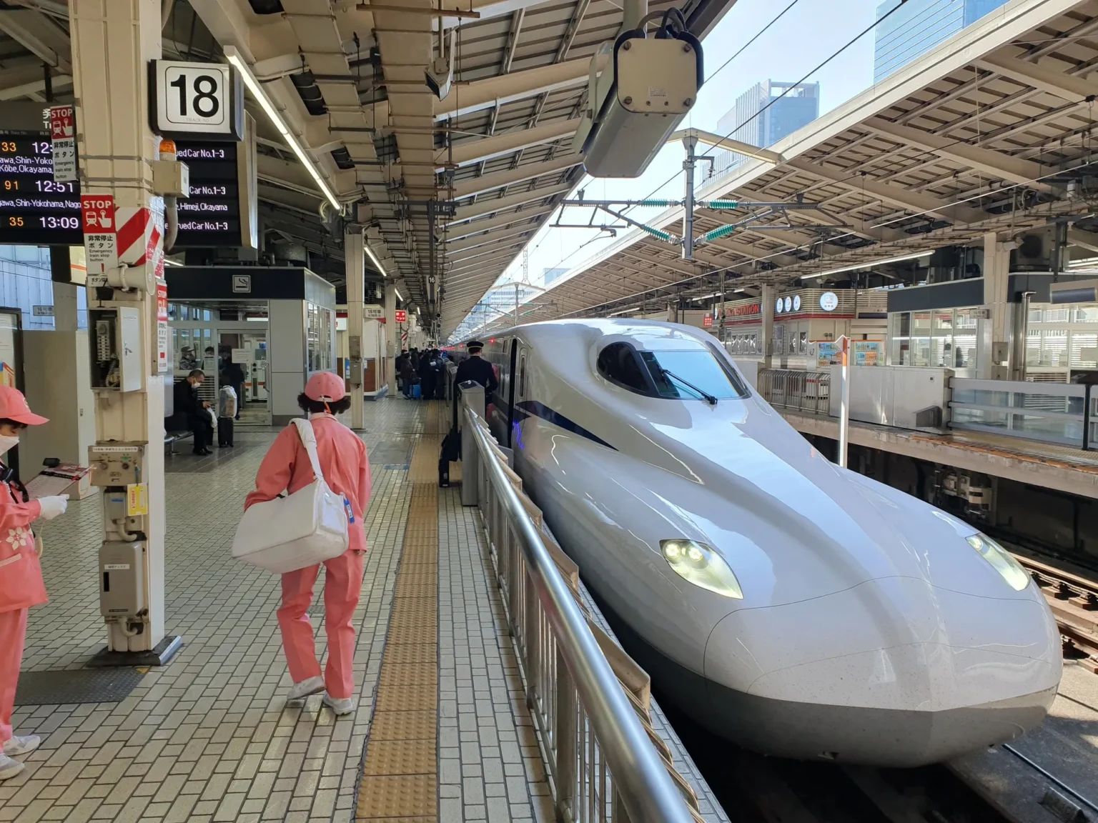The long white nose of an N700S Shinkansen at platform 18 of Tokyo Station, with cleaning staff in pink uniforms and a conductor on the platform and a departure board above