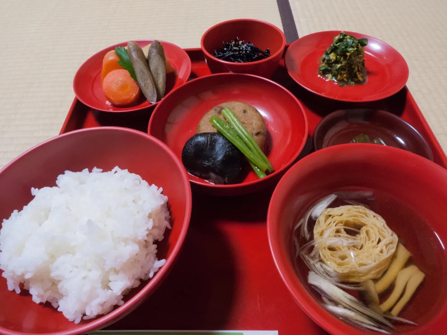 A shojin ryori meal in red lacquer bowls on a red tray: white rice, a clear soup with yuba, simmered vegetables, and small side dishes
