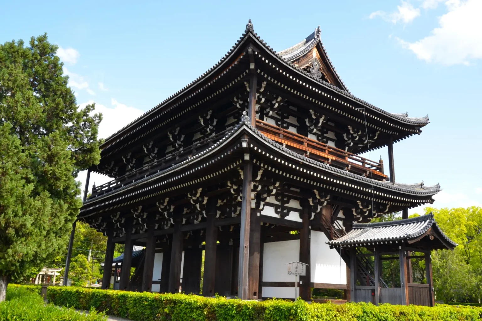 The Sanmon of Tofukuji, a two-storey dark wooden gate with deep tiled roofs, behind a clipped hedge under a blue sky