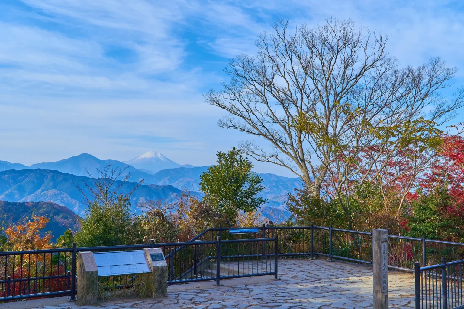 The paved summit viewing area of Mount Takao in western Tokyo, with a wooden post naming the Omiharashi area, red and orange autumn leaves along the railing and snow-capped Mount Fuji beyond the ridges