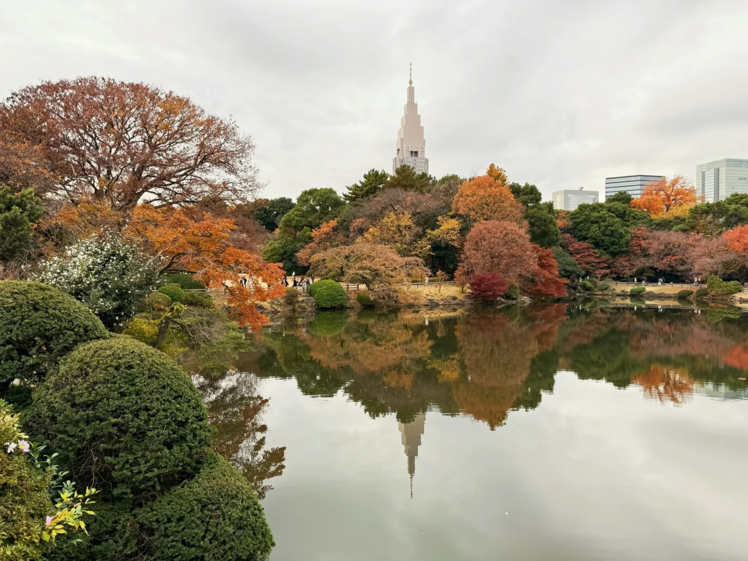 Orange, red and yellow autumn trees around a still pond at Shinjuku Gyoen in Tokyo, with the spire of the NTT Docomo Yoyogi Building rising behind and reflected in the water under a grey sky