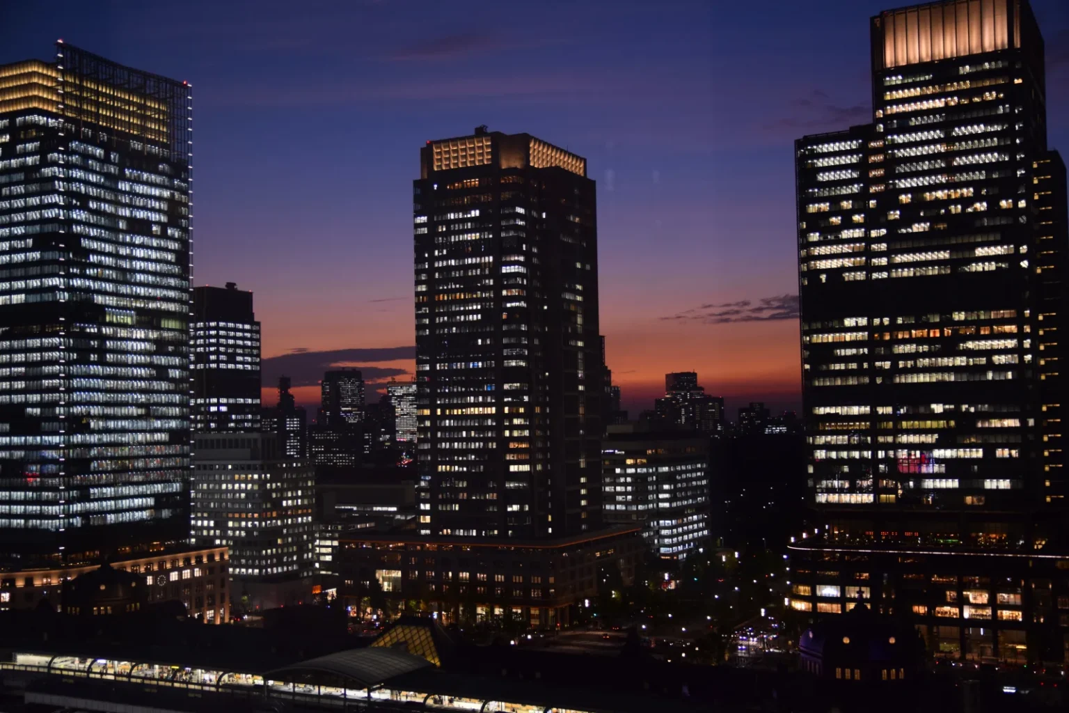 Office towers in Marunouchi with lit windows against a dark blue sky and an orange band of afterglow on the horizon, with lit low buildings and platform roofs in the foreground