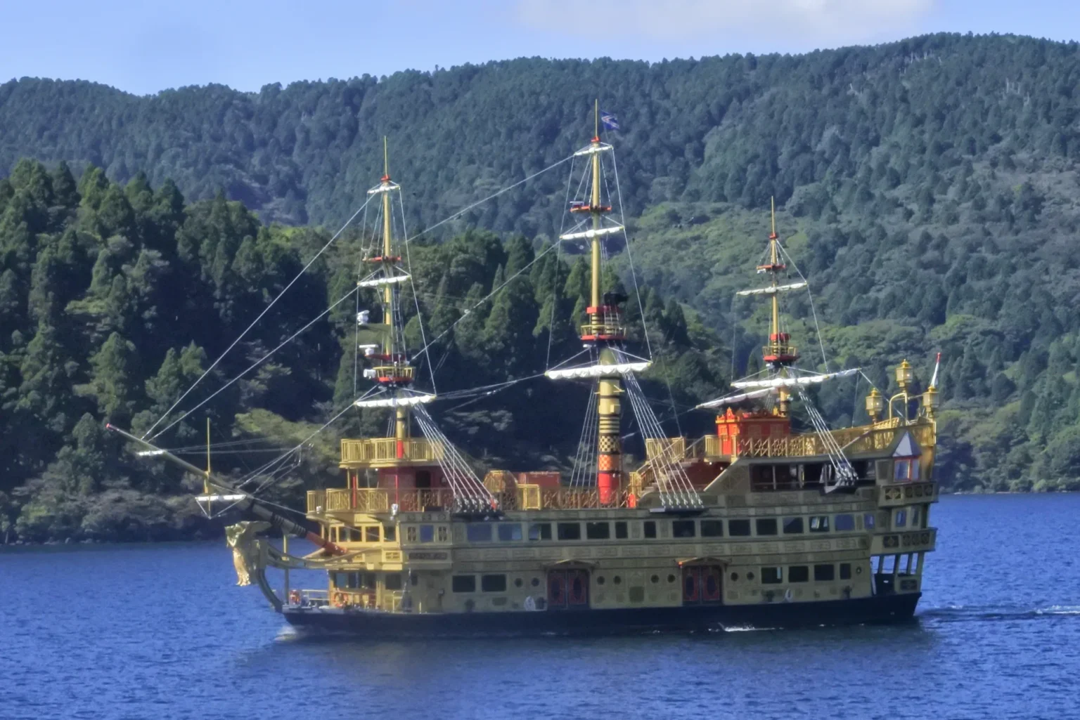 The gold sightseeing ship Queen Ashinoko, styled like a three-masted sailing warship, cruising on blue Lake Ashi in Hakone in front of a steep forested hillside.