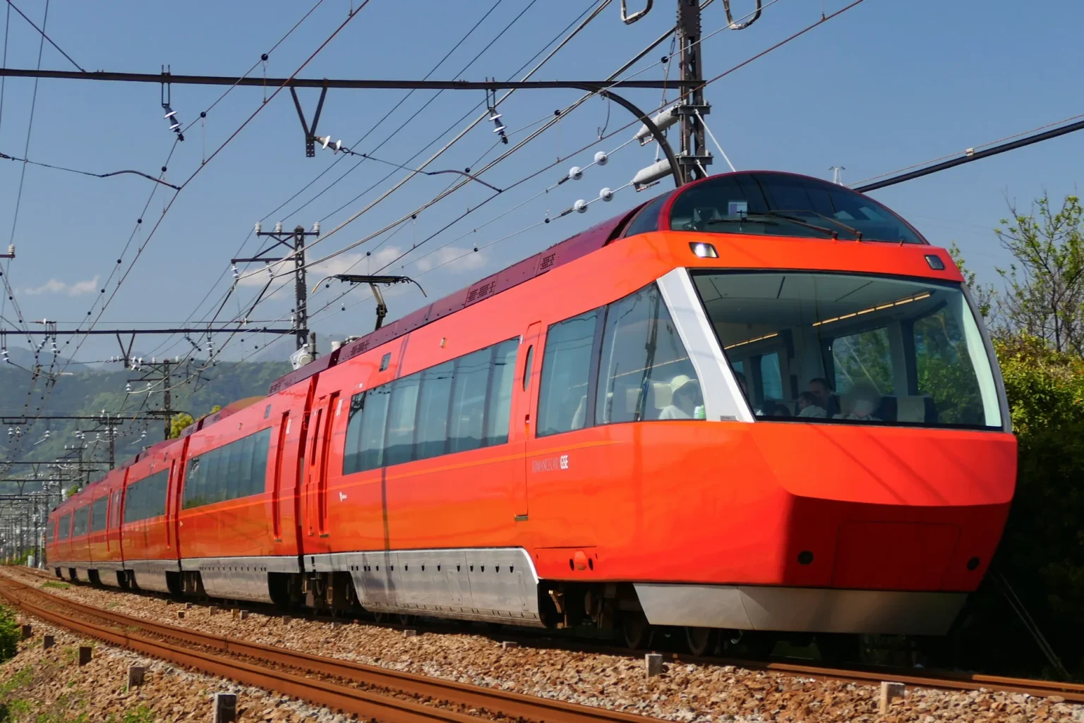 An orange Odakyu 70000-series Romancecar with a raised front observation cab running on ballasted track under overhead wires, with green hills behind, on the Odakyu Odawara Line.