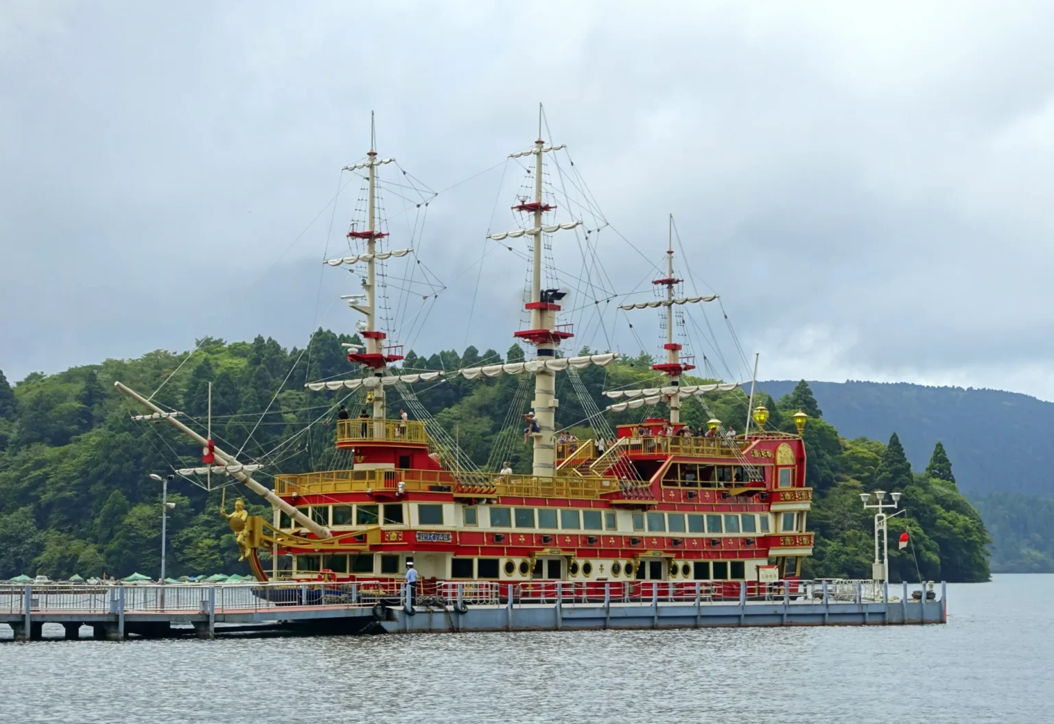 The red and gold sightseeing ship Royal II, built to look like an 18th-century sailing warship with three masts, moored at the Moto-Hakone pier on Lake Ashi under a grey sky, with forested hills behind.