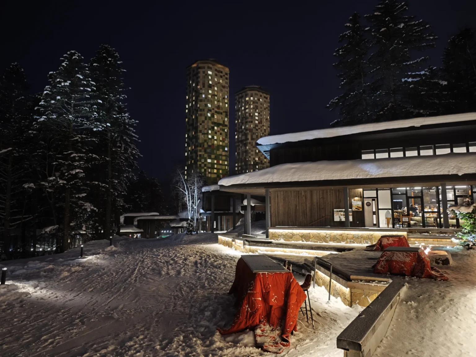 Night in the snow at Hotalu Street, Hoshino Resorts Tomamu: a lit wooden building with a snow-covered roof, high tables wrapped in red quilted covers, a fire pit glowing by stone steps, snow-laden conifers and two tall lit hotel towers against a dark sky, at 17:41 on 14 February 2023