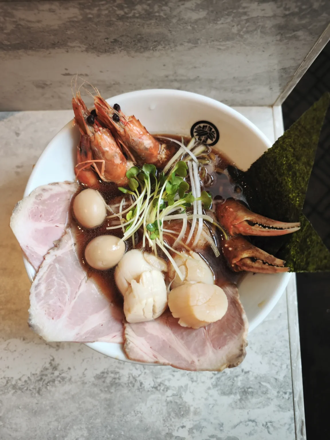 A white bowl of seafood ramen seen from above, with whole prawns, two crab claws, scallops, quail eggs, slices of pork, sprouts and two sheets of nori in a dark soy broth, on a grey stone-effect table at Hoshino Resorts Tomamu at 13:34 on 16 February 2023