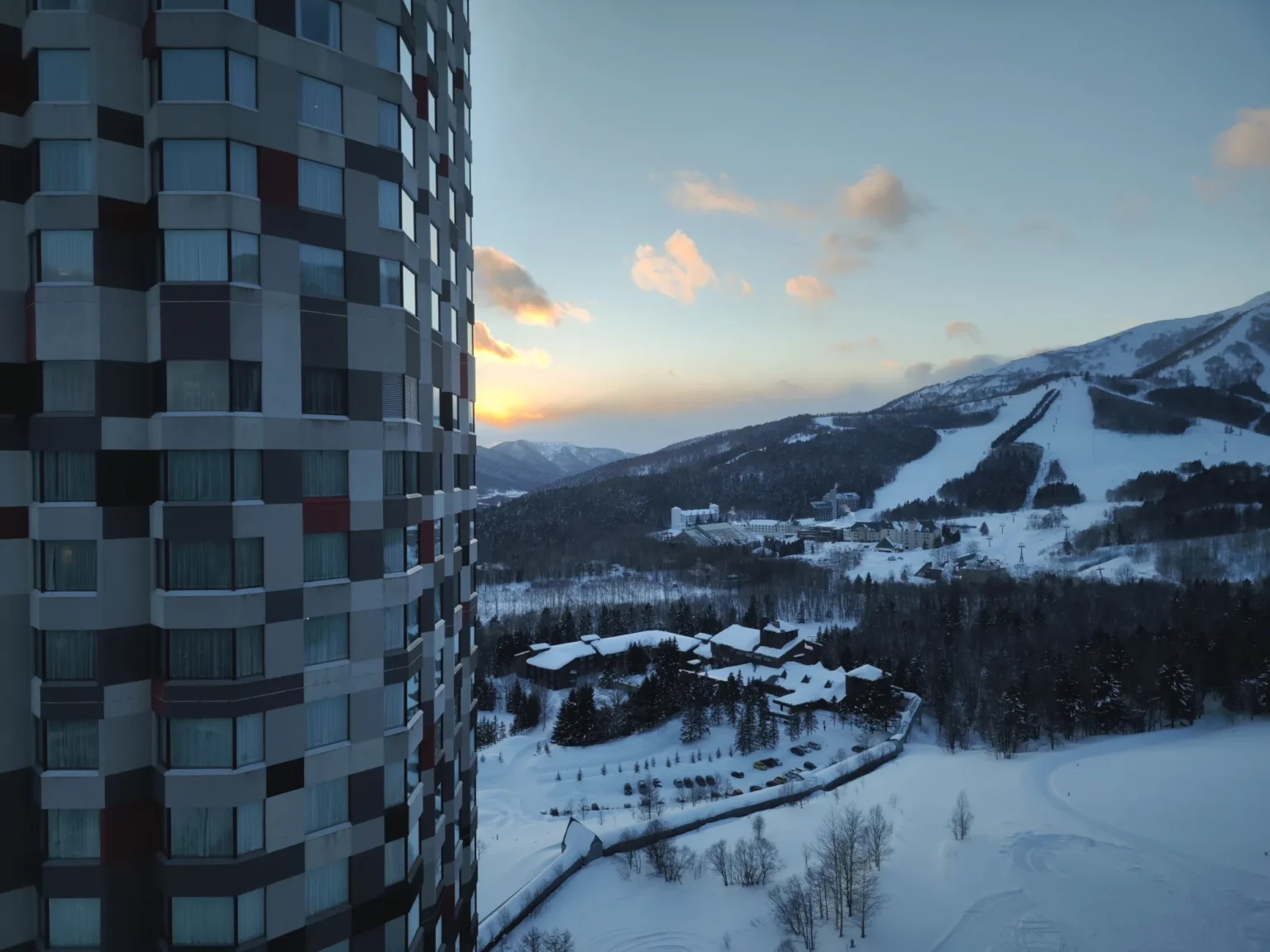 The checked facade of a neighbouring hotel tower on the left, and to the right a snowy valley with low resort buildings, a car park and ski runs on a mountain under an orange sunset sky with pink-lit clouds, seen from a high window at Hoshino Resorts Tomamu at 16:47 on 15 February 2023