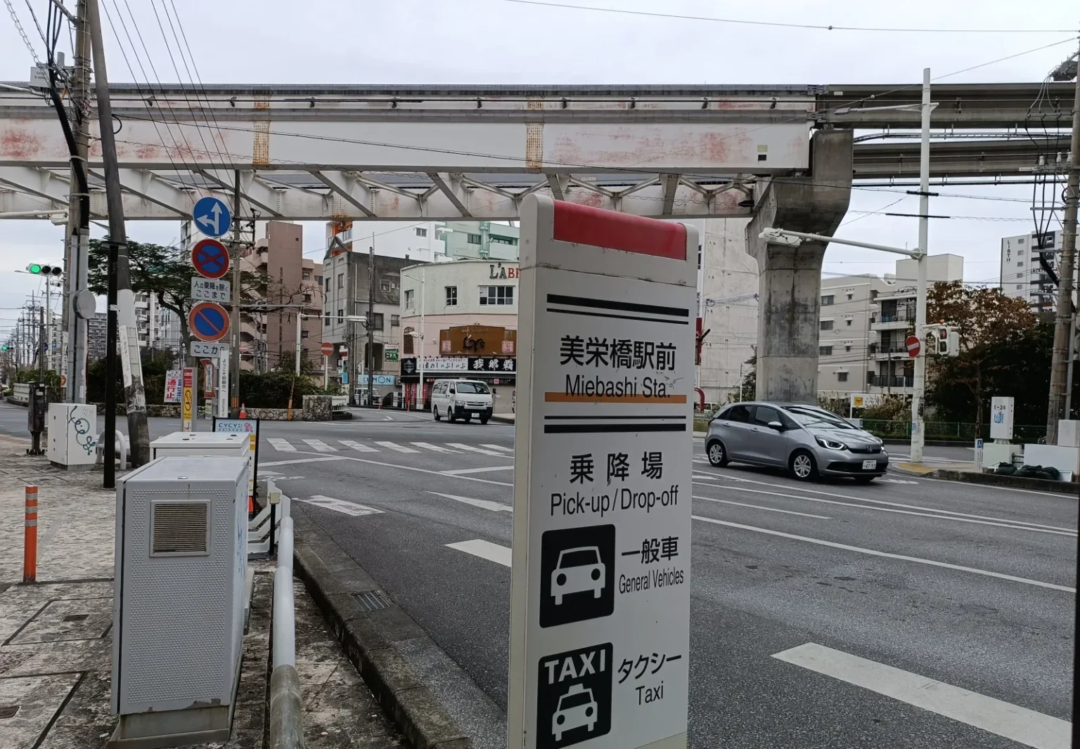 A roadside sign in front of Miebashi monorail station in Naha marking a pick-up and drop-off point for general vehicles and taxis, in Japanese and English, with the monorail track overhead