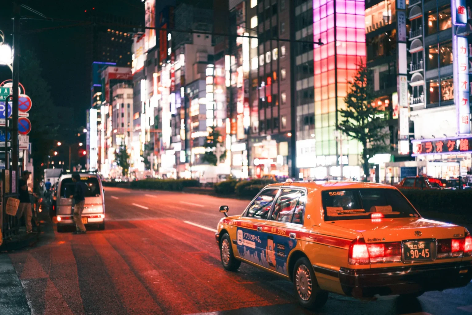 An orange sedan taxi, its side wrapped with an ad reading “call by app” in Japanese, on a wide street in Shinjuku at night, with neon-lit buildings and a white van further up the street