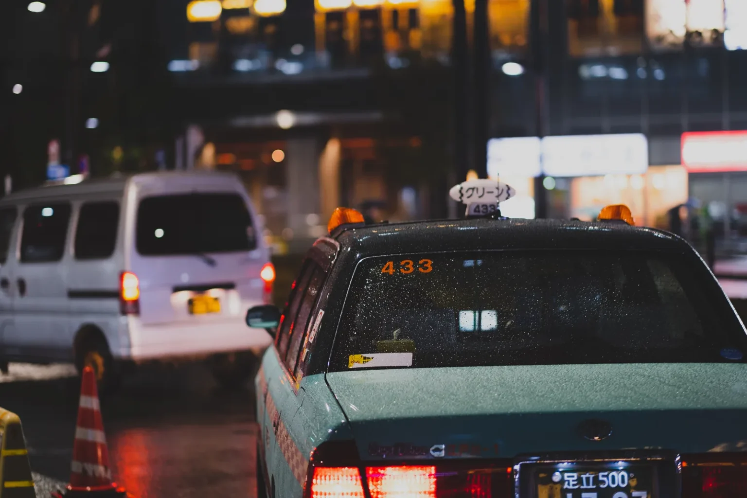 The rear of a green taxi with a lit roof sign and a white van ahead of it on a street near Minami-Senju Station in Tokyo, with lights and a traffic cone in the background