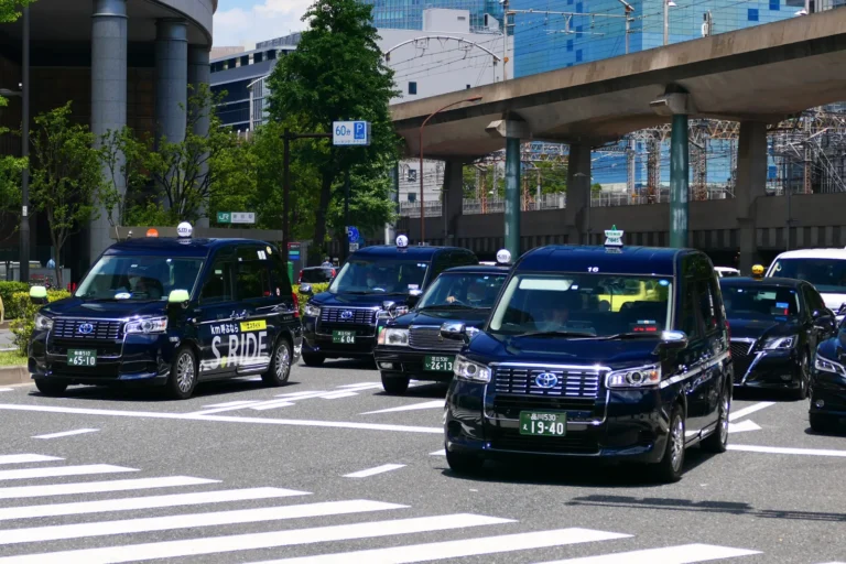 Dark navy Toyota JPN Taxi cabs and a black sedan taxi driving across a zebra crossing near the Marunouchi side of Tokyo Station, under an elevated road, one cab carrying an S.RIDE advertisement on its side