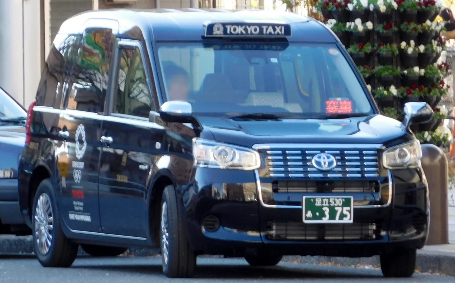 A dark navy Toyota JPN Taxi with a Tokyo Taxi roof sign parked at the kerb, a hanging flower display behind it