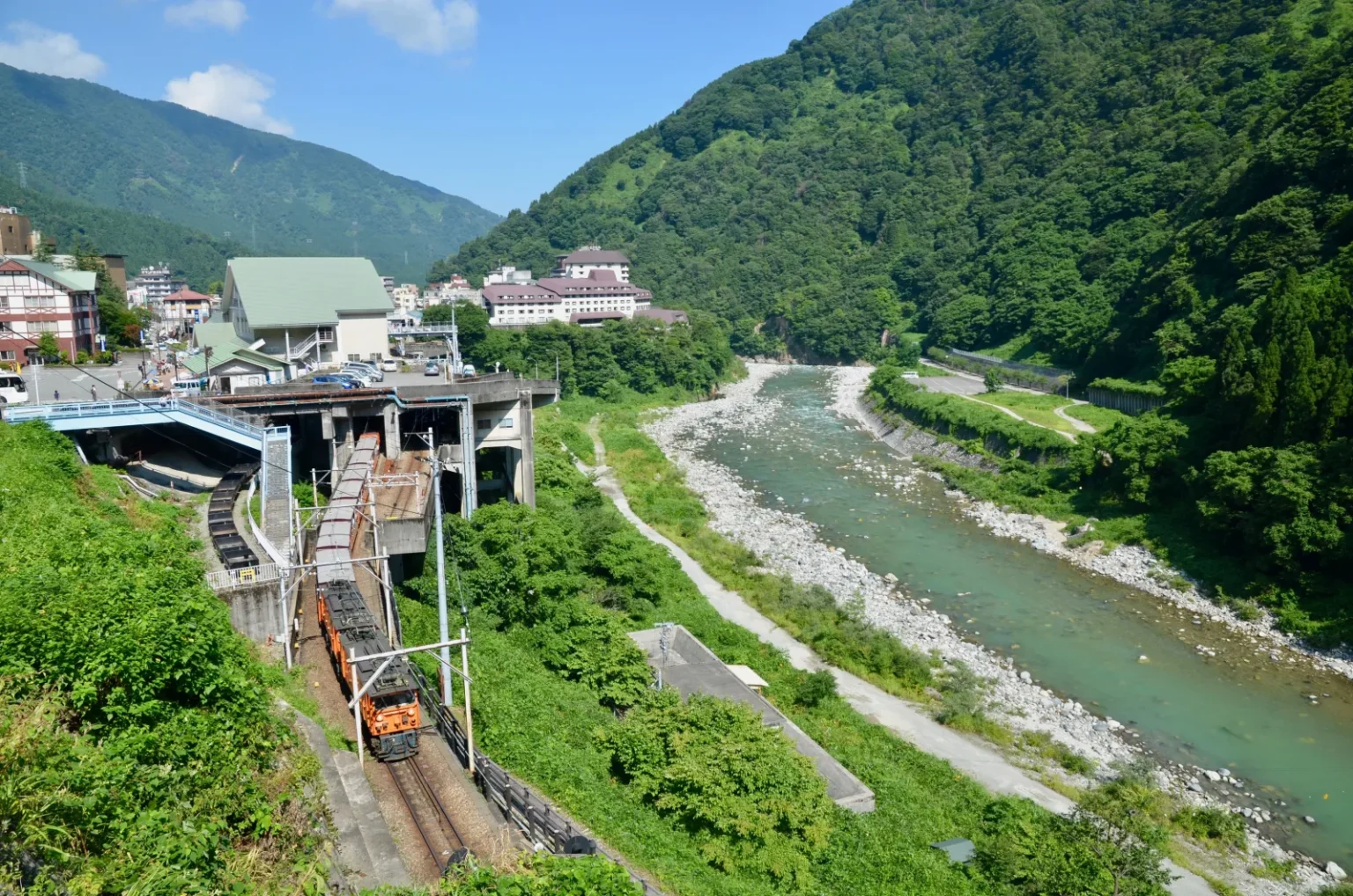 A small orange train on a track below a station building, with hotels of Unazuki Onsen on the hillside and the wide Kurobe River in a green valley