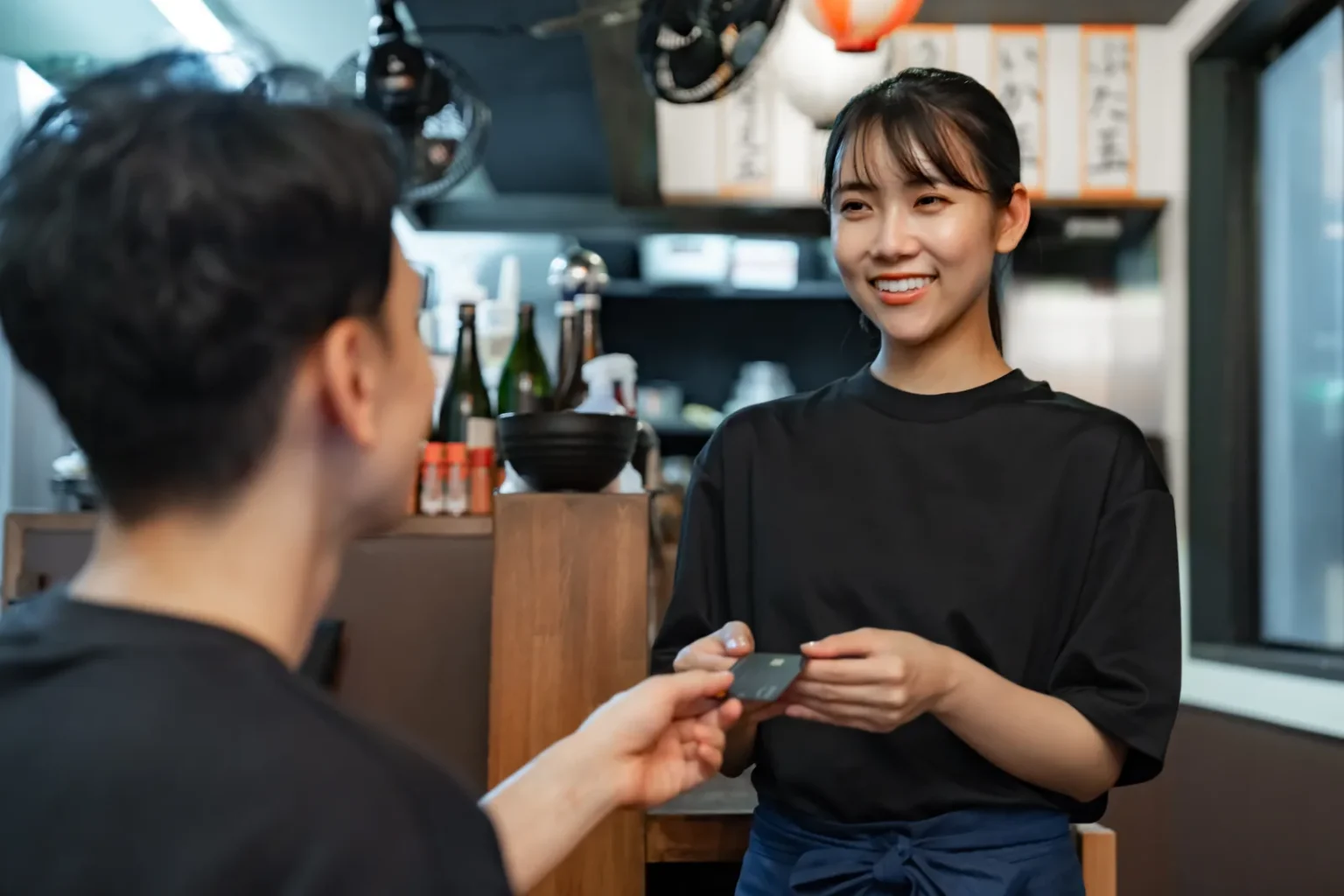 A customer handing a payment card across the counter to a smiling staff member in a black T-shirt and apron at a small Japanese restaurant, with bottles and a bowl on the shelf behind