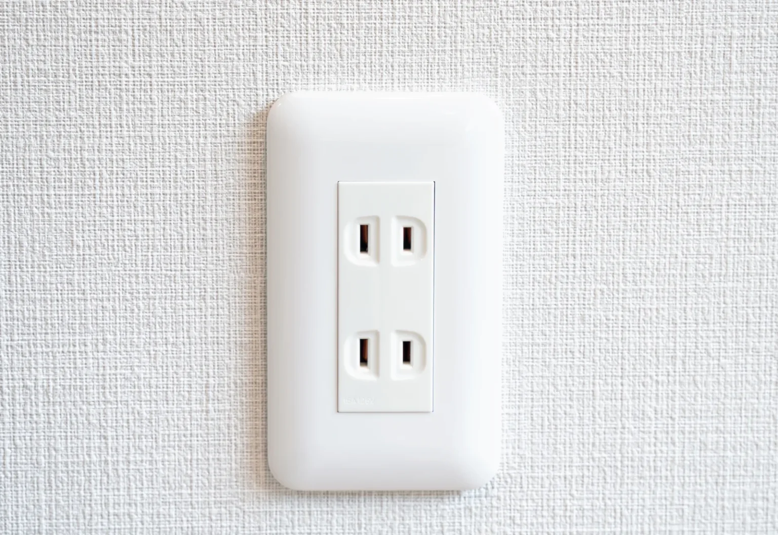 A white double wall socket on a textured white wall, each outlet with two flat parallel slots and no earth hole