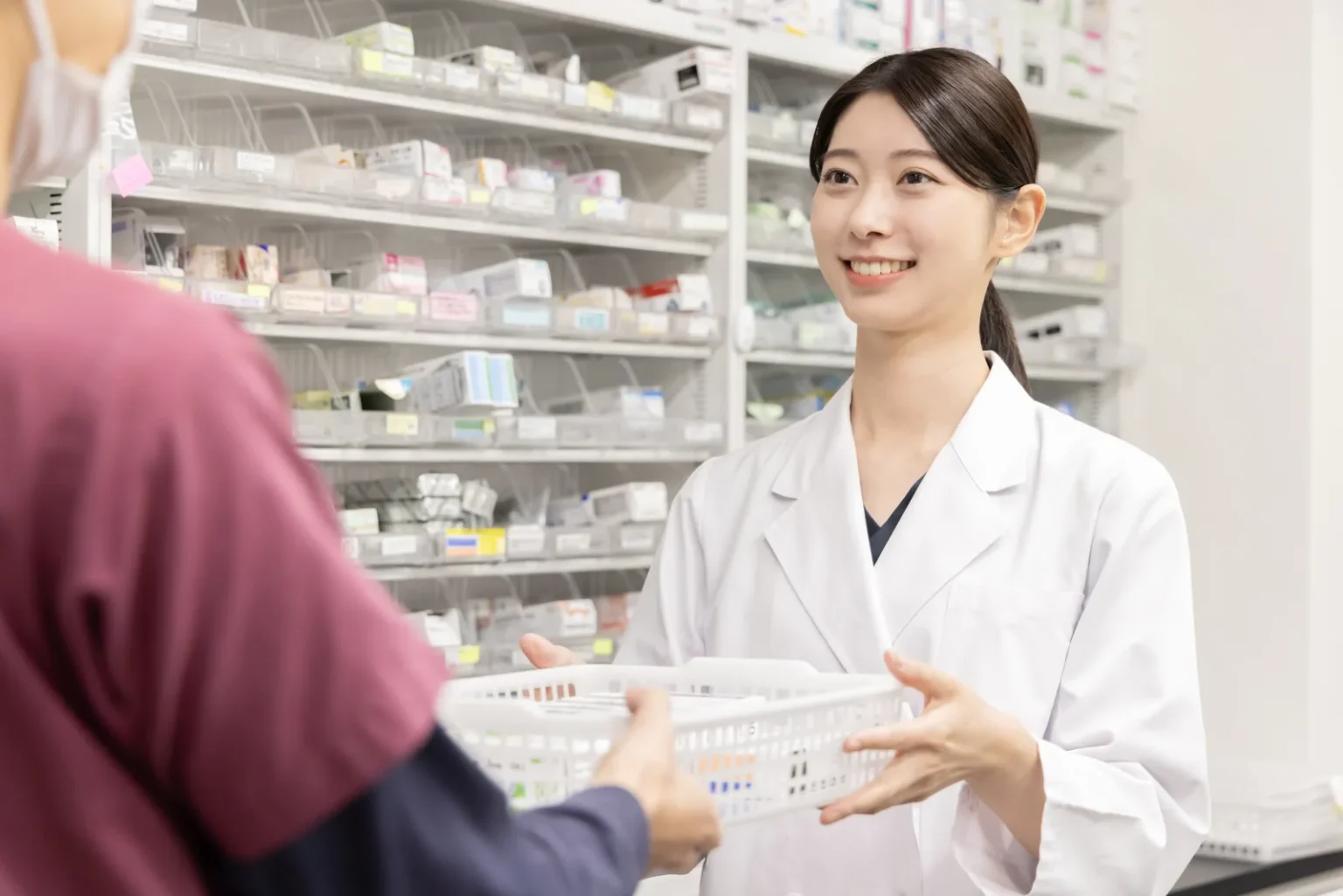 A pharmacist in a white coat smiling and taking a white basket across a counter from a customer, with shelves of medicine boxes in labelled trays behind