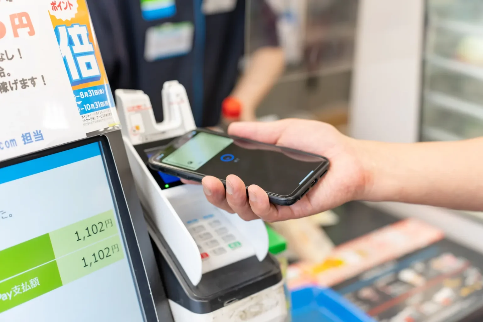 A hand holding a smartphone over the contactless reader of a Japanese convenience store till, with the amount 1,102 yen on the customer display screen