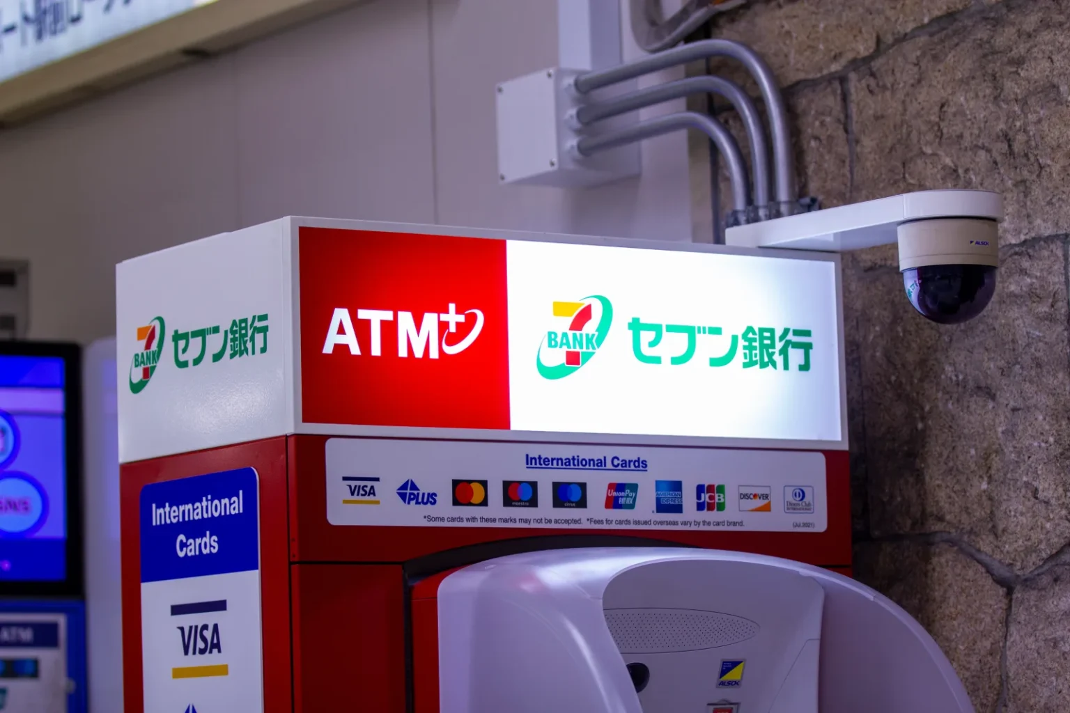 A Seven Bank ATM with a red and white illuminated sign, an International Cards panel showing card logos including Visa, Mastercard, UnionPay, American Express, JCB and Discover, and a security camera on the wall beside it