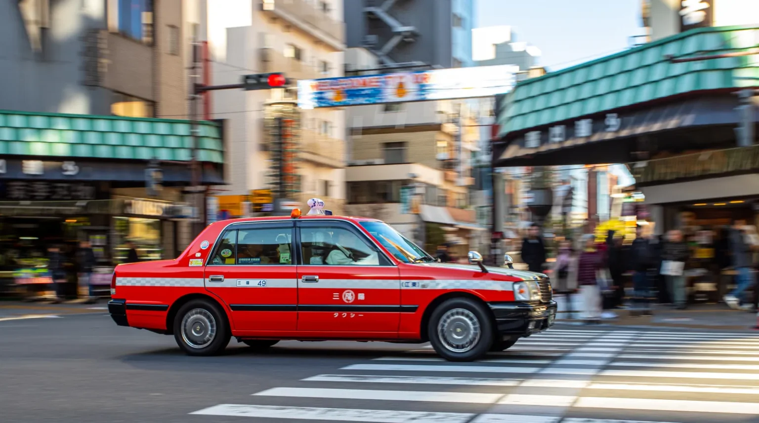 A red and white Japanese taxi with a roof light driving past a pedestrian crossing in a shopping street, with shoppers and green-roofed shop fronts behind it