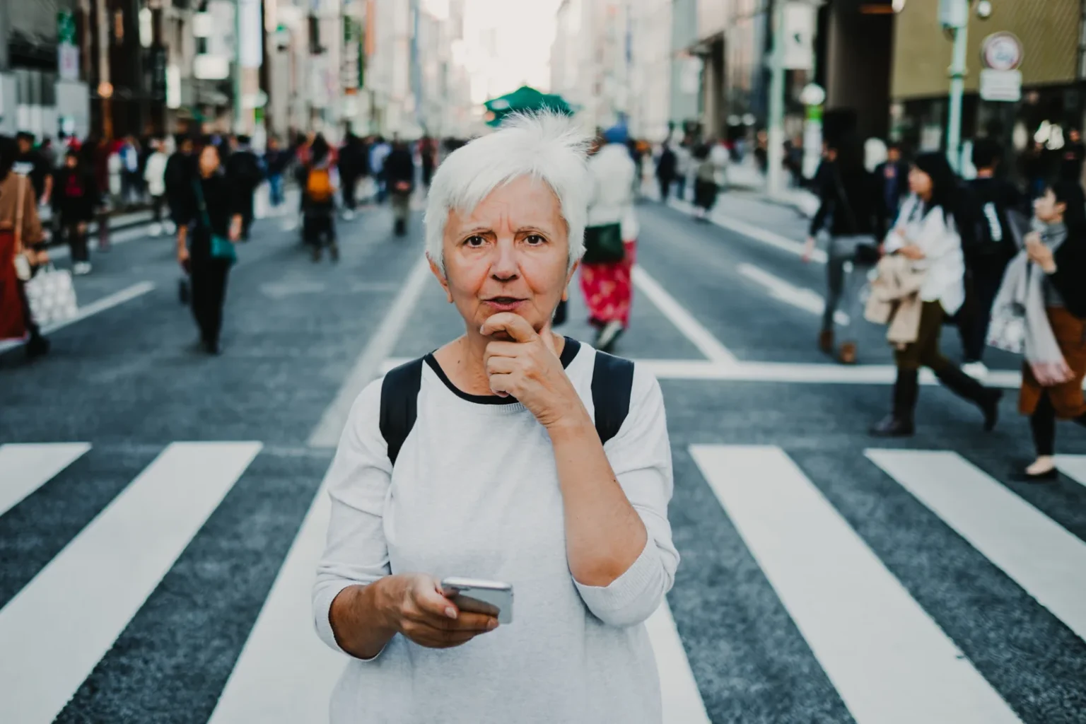 A traveller with short white hair and a backpack standing on a pedestrian crossing in a busy shopping street, holding a smartphone in one hand and resting their chin on the other